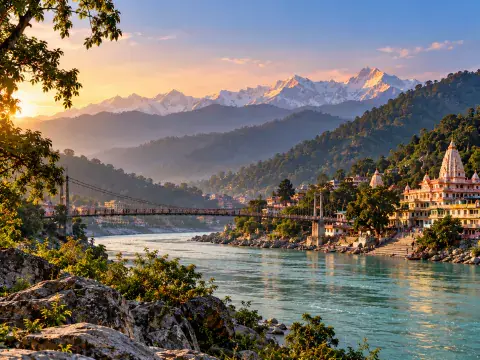 Suspension bridge across a wide turquoise mountain river with riverside temples and snow-capped Himalayan peaks in the background at sunrise.