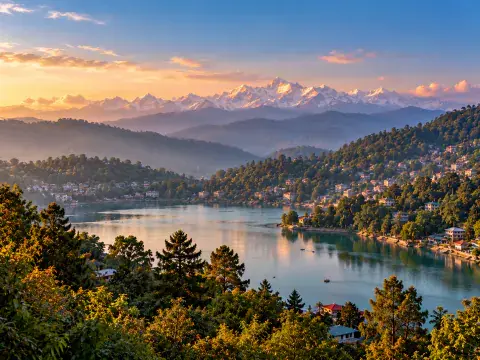 Scenic hill station lake surrounded by forested slopes with snow-capped Himalayan peaks illuminated at sunset.