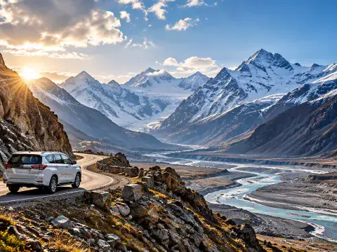 White vehicle driving on a mountain road beside a wide river valley with snow peaks in the distance.