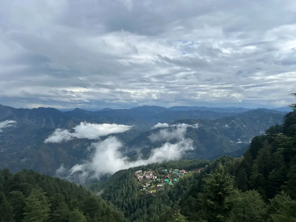 A quiet Himalayan valley with scattered houses surrounded by dense pine forests and mountain slopes under low clouds.