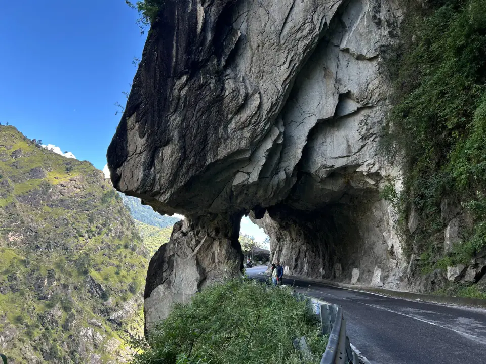 Two travellers standing beside a paved mountain road carved directly underneath a massive rock overhang.