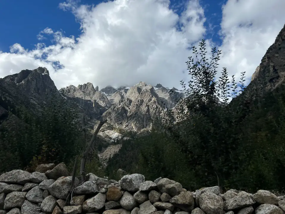 A rustic dry-stone wall bordering a mountain trail with rugged granite peaks and drifting clouds in the distance.