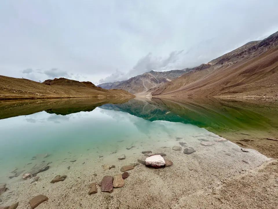 A serene high-altitude glacial lake with crystal-clear emerald water reflecting surrounding barren mountain ridges under a soft overcast sky.