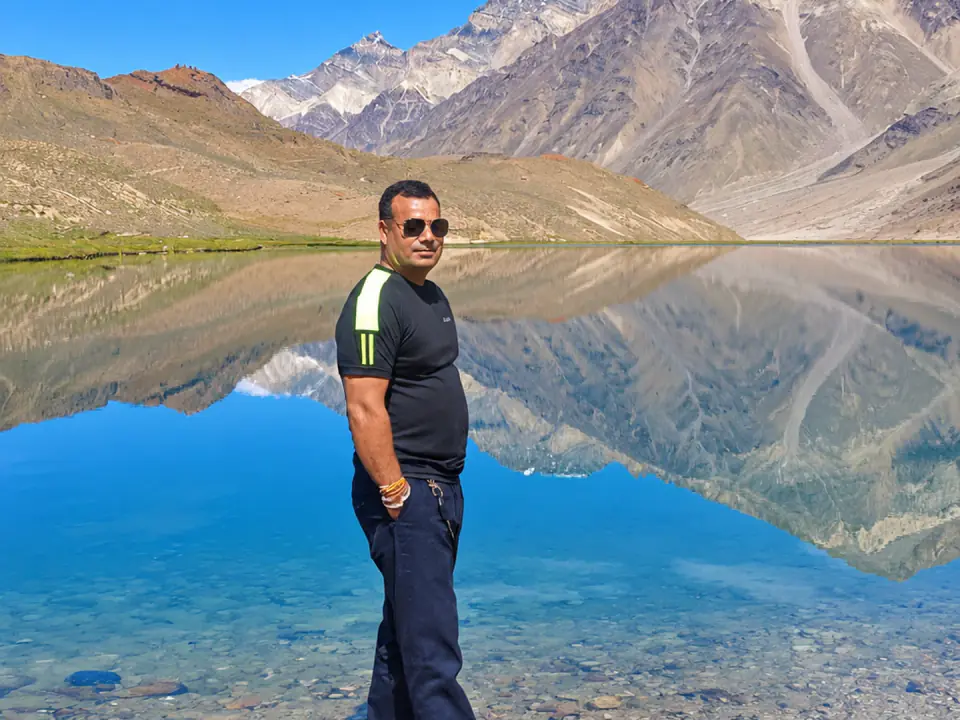 Bhupinder standing beside the turquoise waters of a tranquil mountain lake with rugged Himalayan peaks reflecting in the surface.