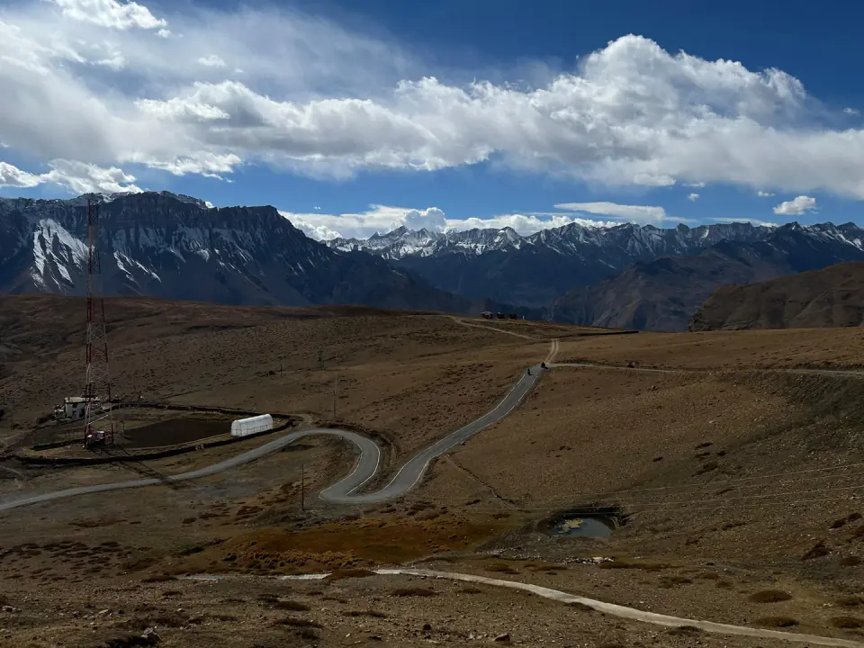 A winding mountain road sweeping through a high-altitude Himalayan valley plateau against rugged snow-capped peaks under a partly cloudy sky.