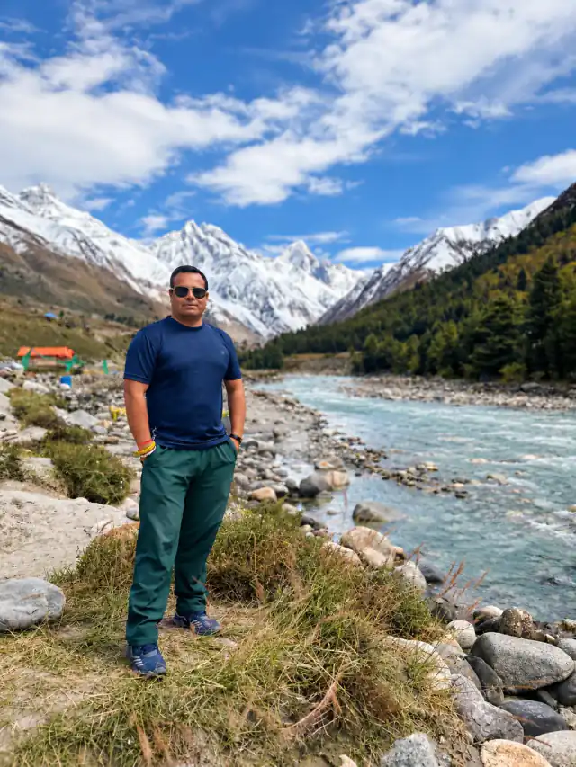 Man standing on a rocky riverbank in front of a turquoise mountain river and snow-capped peaks under a blue sky